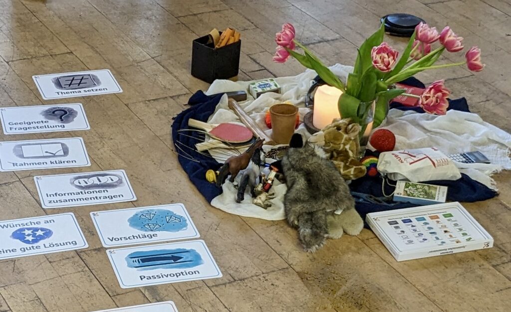 Auf dem Boden liegen Karten mit Schritten des Systeminen konsensieren. Daneben Blumen, Gegenstände und Kerze auf einem Blauen Tuch gestaltet.